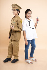 Happy young indian girl child wearing doctor uniform holding stethoscope and boy kid wearing police costume dress isolated on beige studio background. education concept