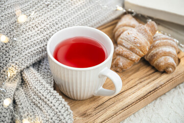 A cup of red tea, a croissant and a knitted element on a wooden tray.