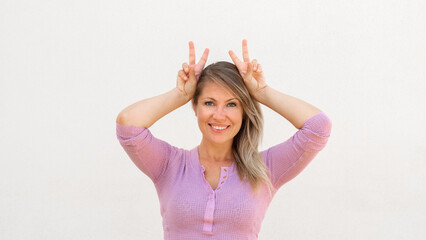 Happy Caucasian woman mimicking bunny ears. Portrait of cheerful mature female model with fair hair...