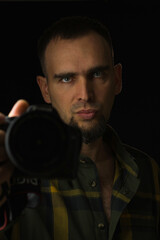 Breast portrait of photographer on black background. Bearded man in green plaid shirt holding camera in outstretched hand and looking into camera. Low key.