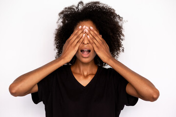 Close-up of excited young woman covering eyes with hands over white background. Portrait of African American lady wearing black T-shirt and jeans standing in amazement. Surprise concept