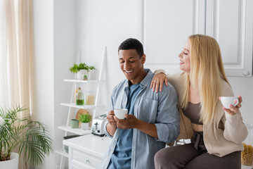 Obraz na płótnie Canvas joyful blonde woman sitting on kitchen worktop near african american boyfriend with coffee cup