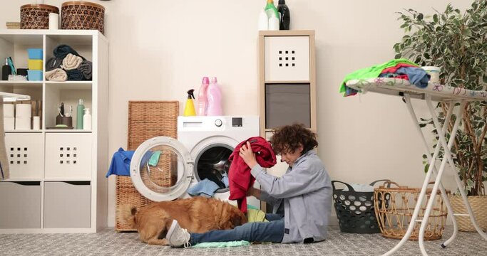 Housework. Boy doing laundry at home loading clothes into washing machin and playing with dog.