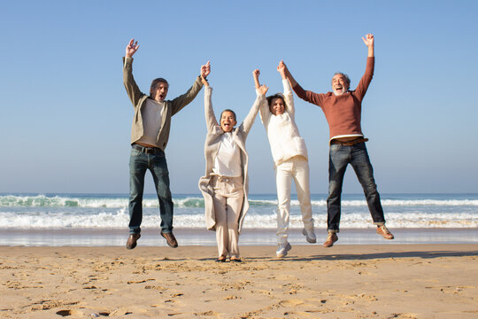 Group of joyful senior friends having fun at beach on sunny autumn day. Four people laughing and jumping up high while raising their arms in triumph. Vacation, joy, friendship, retirement concept