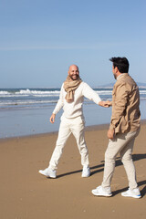 Carefree gays walking together on beach. Positive bald man in scarf holding hand of boyfriend and looking at him. Stroll concept