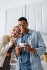 smiling multiethnic couple holding morning coffee in kitchen