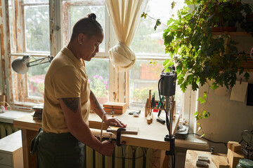 Side view portrait of tattooed female artist cutting wood while standing by window in cozy workshop, copy space