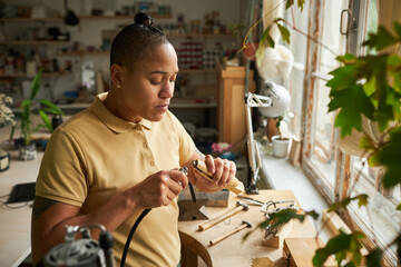 Portrait of black female artist working on handmade jewelry in cozy workshop lit by sunlight