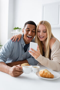 Excited Interracial Couple Looking At Mobile Phone And Laughing During Breakfast