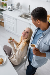 high angle view of blonde woman looking at african american boyfriend while drinking morning coffee in kitchen