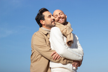 Happy carefree gays embracing outdoors. Affectionate brunette man whispering in ear of bald boyfriend in scarf against blue sky. Love concept