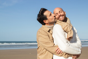 Obraz premium Bearded man kissing boyfriend on beach. Brunette middle-aged man with closed eyes hugging partner from back. Gays concept