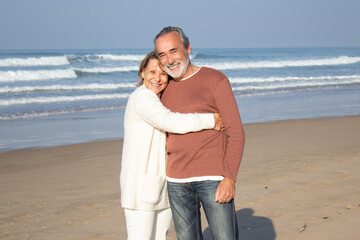 Beautiful senior couple standing at seashore on sunny day, smiling and looking at camera. Happy blonde woman hugging gray-haired bearded man, showing affection. Love, holiday, retirement concept