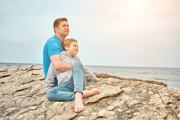 Happy family, father and son bonding, sitting on stone by the sea looking at view enjoying summer vacation. Togetherness Friendly concept
