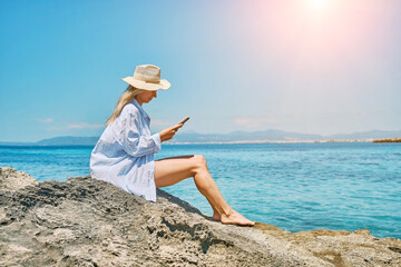 Fashion pretty woman outdoors lifestyle watching, reading on tablet ebook on the beach in summer day. Wearing wide brimmed hat, with uv protection. Concept of beach vacation.
