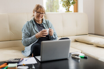 Serene mature female with blond hair having hot tea or coffee while working remotely in front of laptop at home