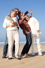 Group of middle-aged friends having fun at the beach, standing against sea background and taking selfie. Happy man holding smartphone and making shot. Modern technology, friendship, leisure concept