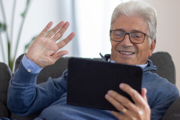 Smiling elderly man videoconferencing with family using digital tablet and waving hand. Positive gray-haired man sitting on sofa at home. Online chatting concept