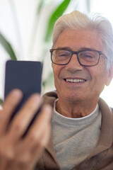 Happy aged man chatting via smartphone app. Smiling handsome man in eyeglasses calling via video chat at home. Wireless technology concept