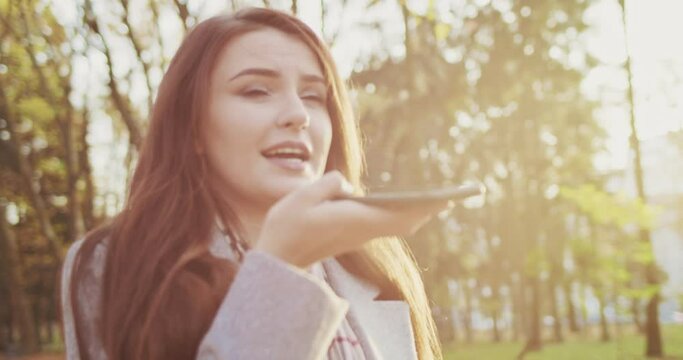 Close up of young charming Caucasian woman strolling in park and recording voice message by smartphone. Outside. stylish female having walk on fall day and sending audio talk on mobile phone.
