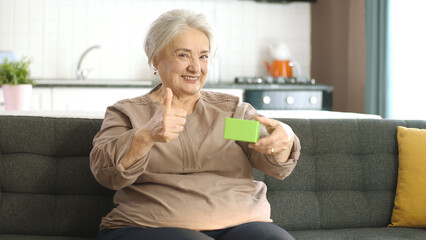 The old woman is holding something green in her armchair, showing a product, smiling and presenting a cheerful,imaginary object.Creative 3d artists can replace the green box with any product they want