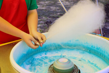 A portion of cotton candy. Hands hold a stick for making dessert. Rest with family in the amusement park.