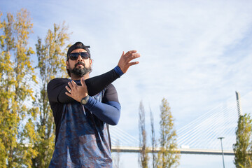 Mid adult man in sunglasses getting ready for jog. Man in sport clothes stretching arms in park on summer day. Sport, training concept