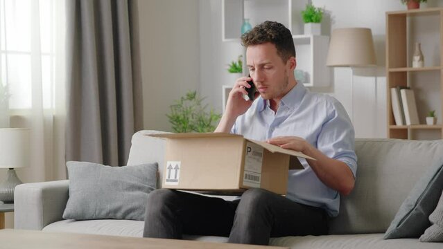 Man sitting on sofa with opened parcel express complaints to seller or courier on the phone about damaged goods, crashed items inside cardboard box
