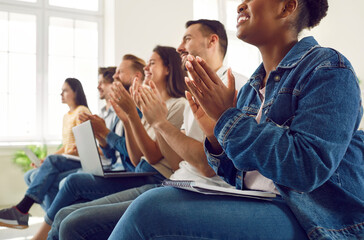 Satisfied diverse audience of students or workers applauding during lecture or seminar. Cropped image of multiracial people in denim casual clothes sitting in row on chairs with notebooks and laptops.