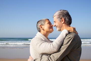 Romantic senior couple hugging at seashore on sunny autumn day. Short-haired middle-aged lady kissing her husband tenderly while spouses enjoying vacation at seaside. Side view. Travel, love concept