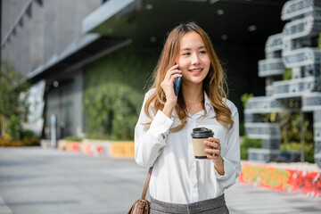 Fototapeta premium Beautiful woman walking outside in street and calling talking on mobile phone outdoors near office, portrait lifestyle businesswoman with bag cell to customer while holding coffee paper cup takeaway
