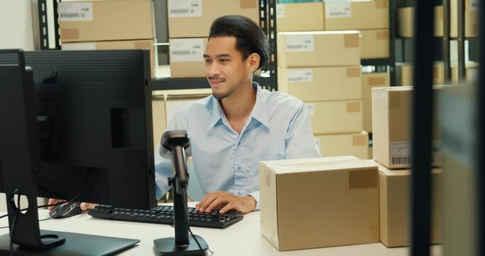 Happy Asian man wear formal uniform shirt sit in front of desk use computer and barcode machine scanner record customer online data in cardboard box delivery at warehouse. Startup business concept.