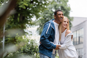 joyful interracial couple in hoodies embracing and looking away on green and blurred street