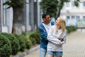 Fototapeta premium joyful african american man embracing young blonde girlfriend on blurred street in city