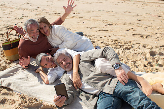 Senior friends taking selfie at beach on sunny day. Two middle-aged couples relaxing at sandy shore while gray-haired man holding smartphone and taking photo. Leisure, modern technology concept