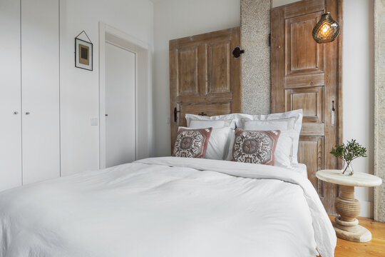 Bright bedroom interior design. Double bed with white linens, white walls and wooden doors as decor