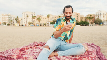 Fototapeta na wymiar Smiling man sitting and relaxing on the beach with smartphone in his hands