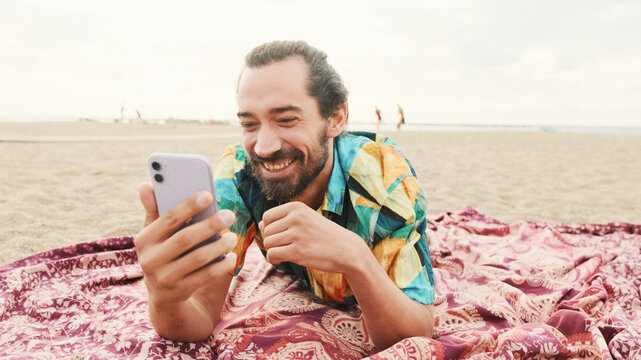 Young happy man lies talking by video call on the beach