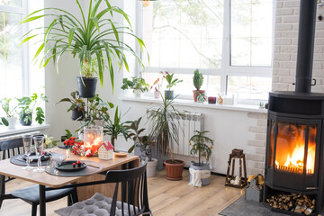 Festive interior of house is decorated for Christmas and New Year in loft style with black stove, fireplace, Christmas tree. Warm studio room with set table, burning wood, cozy and heating of home
