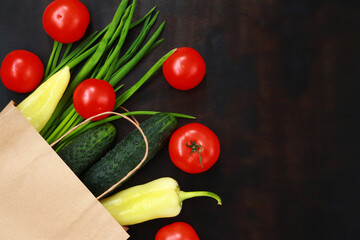 Paper bag with spilled vegetables on a dark wooden table, top view. Ripe tomatoes, green onions, cucumbers and yellow peppers on the table. The concept of healthy eating and vegetarianism