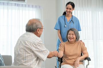 Senior man holding senior woman or his wife patient while sitting on wheelchair. Asian young woman caregiver doctor helps support her during home visit.