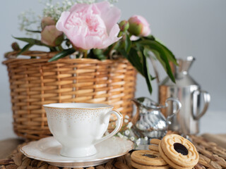 An artistic still life with pink peonies in a wicker basket, a cup of coffee, cream and cookies. Horizontal photo.