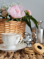 An artistic still life with pink peonies in a wicker basket, a cup of coffee, cream and cookies. Vertical photo.
