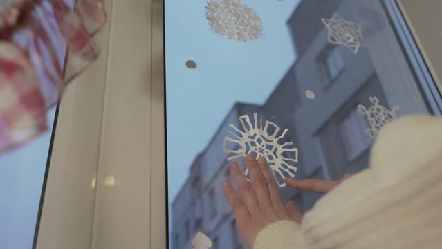 Teenage girl sticking paper white snowflake on window glass in Christmas holiday eve