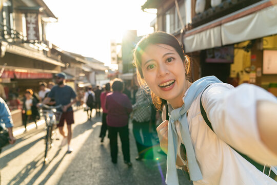 smiling asian Japanese girl visitor looking at camera while taking selfie picture at retro shopping street of Ninenzaka and Sannenzaka in backlight at sunset in Kyoto japan