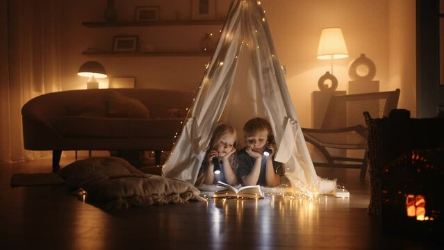 little siblings are playing in handmade wigwam from blanket in living room in winter night