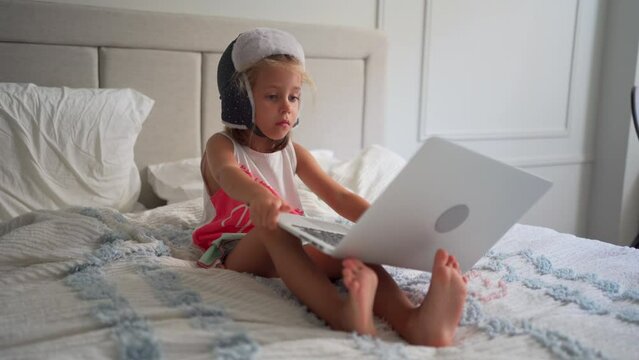 Child in funny hat watching video on laptop sitting sofa at home. Little caucasian girl enjoy cartoons on laptop sitting bed in bedroom. Joyful preschool kid girl use laptop computer