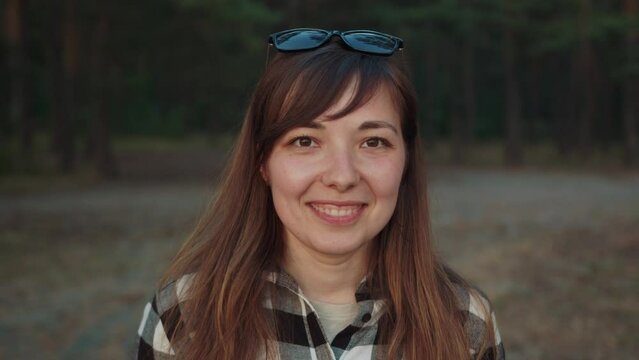 Portrait of charming young girl with glasses on her head and in plaid shirt on the background of the forest. Slow motion