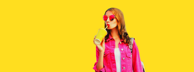 Portrait of stylish young woman drinking juice wearing pink jacket, sunglasses on yellow background