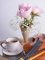 An artistic still life with a bouquet of pink peonies, a cup of tea and a violin. Vertical photo.